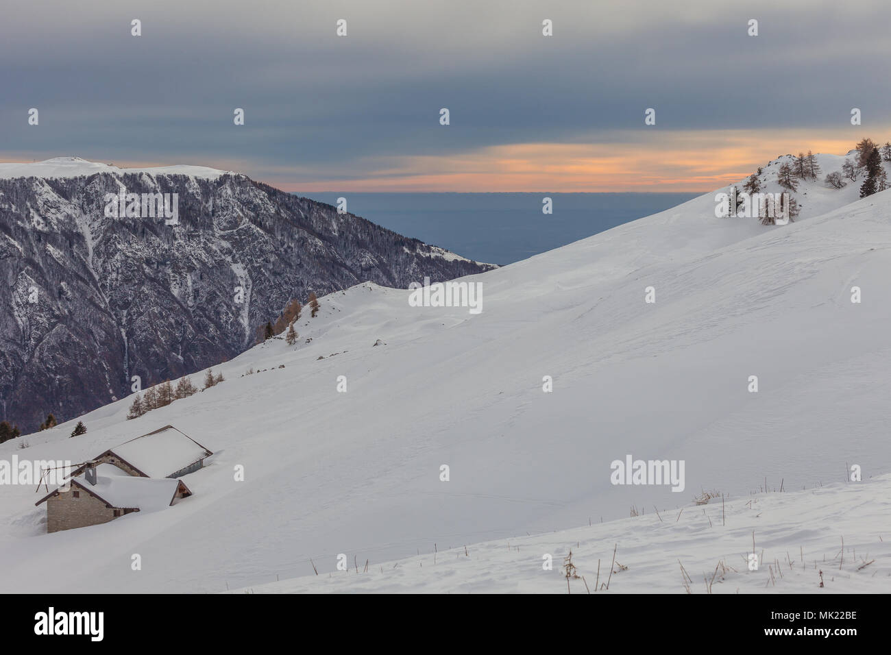 Small mountain farmhouses on snowy slope with a gray sky, a pale sun, mountains and plain in the background, Belluno, Veneto, Italy Stock Photo