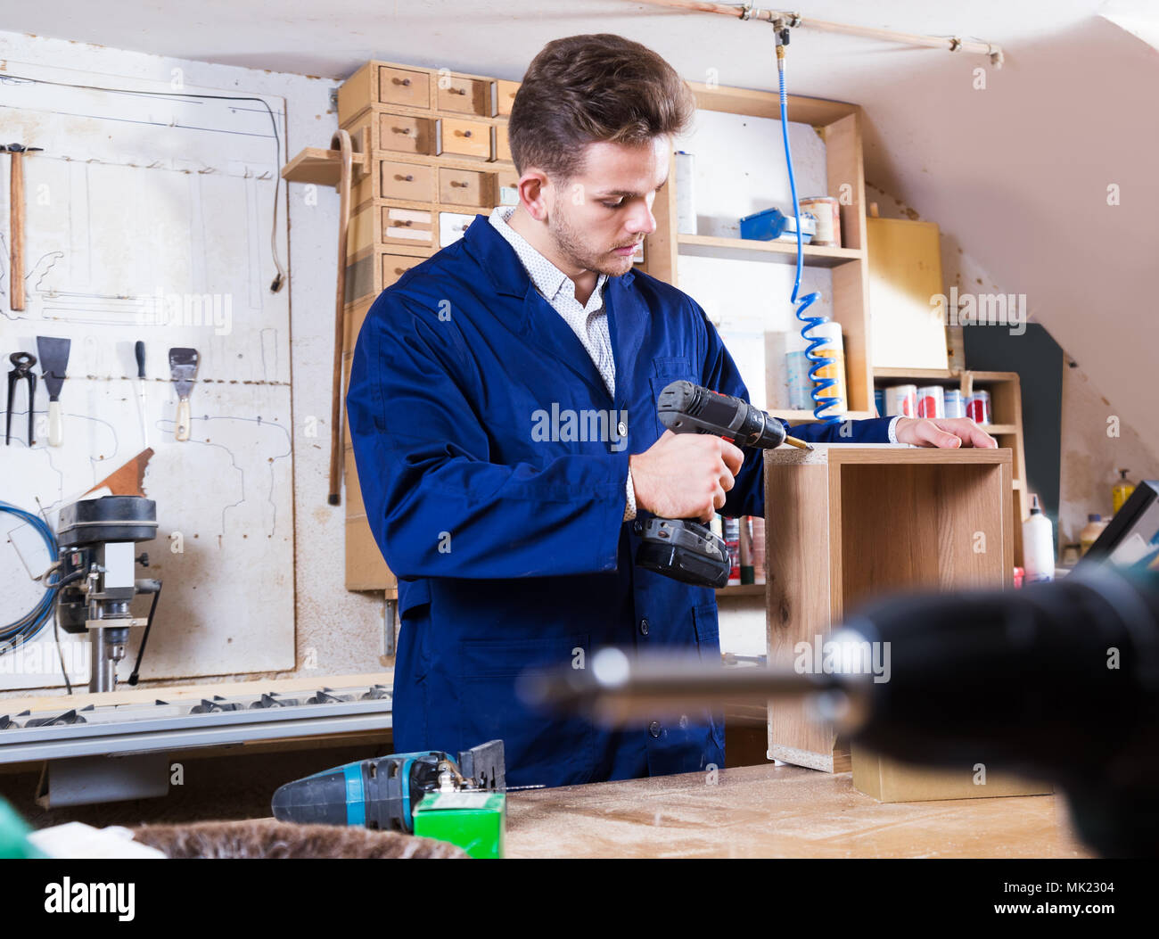 Young smiling man using drilling machine to make drawer at workshop Stock Photo