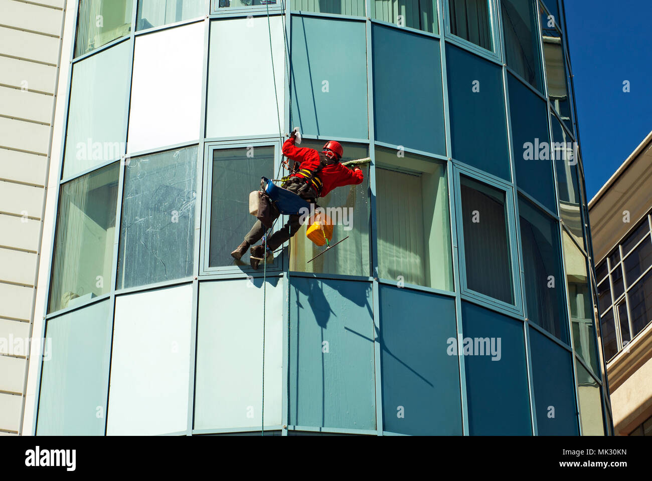 worker in a helmet washes the window of a high-rise building from the outside, hanging on a rope Stock Photo