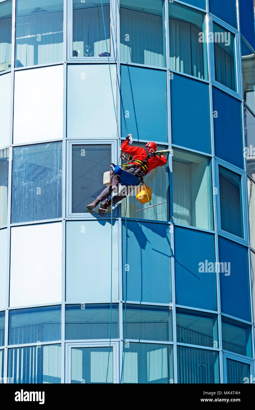 worker in a helmet washes the window of a high-rise building from the outside, hanging on a rope Stock Photo