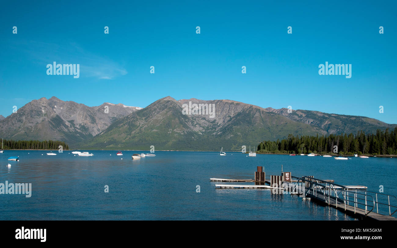 Boat dock on lake of blue water with boats and forest on each side of lake with tall Rocky Mountains beyond under blue sky. Stock Photo