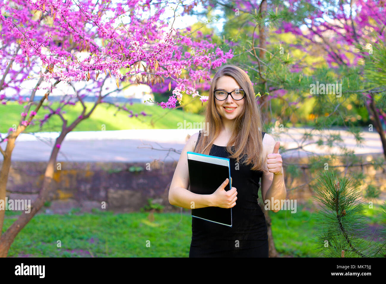 Cheerful female student showing thumbs up in garden with documents in ...