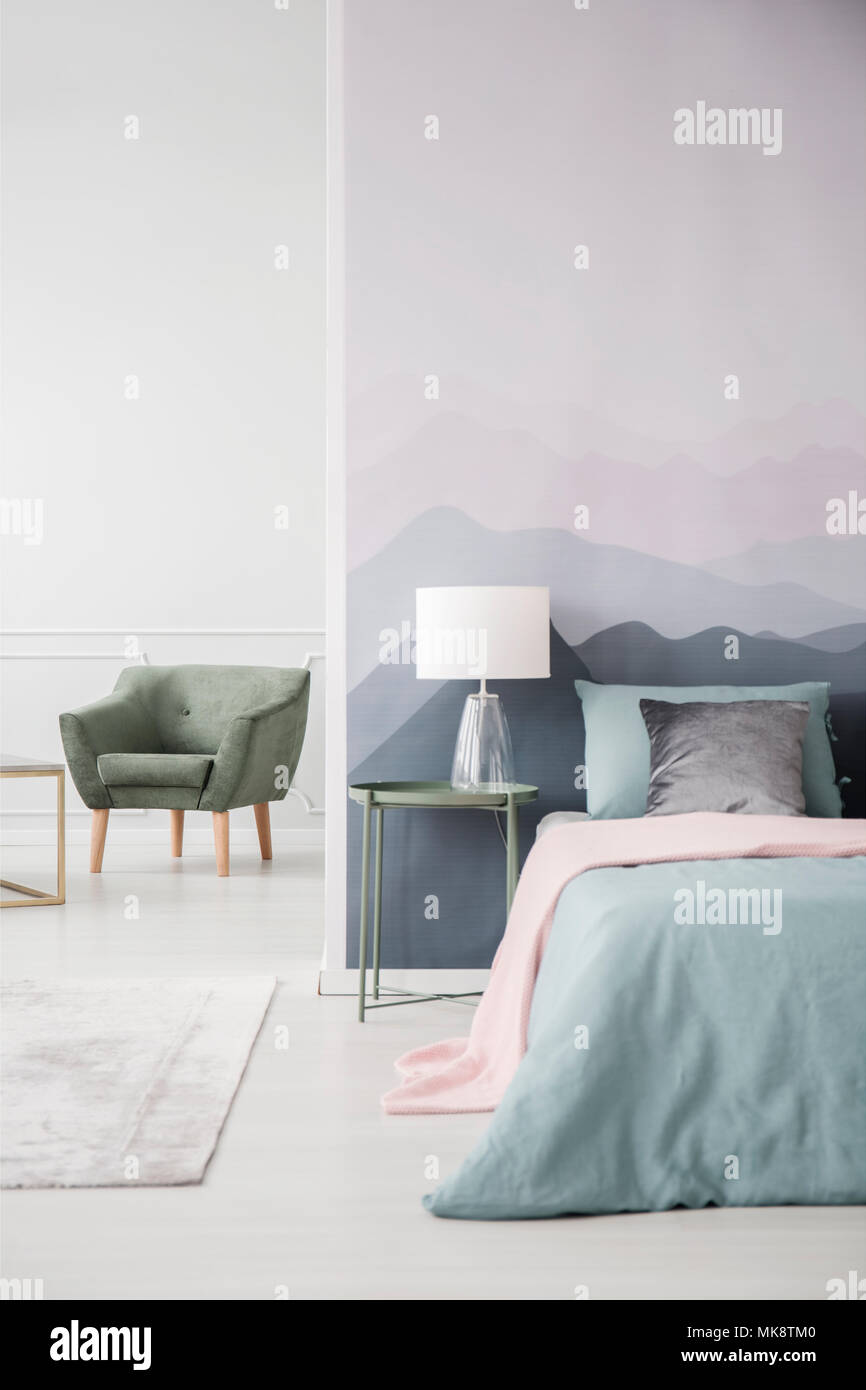 Pink blanket on bed next to a green table with lamp against mountain wall in bedroom interior with armchair Stock Photo