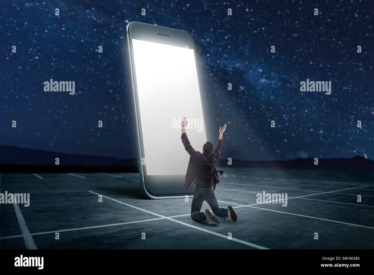 Phone addicted people concept. Man praying on his knees against large smartphone with glowing screen. Scaling effect Stock Photo