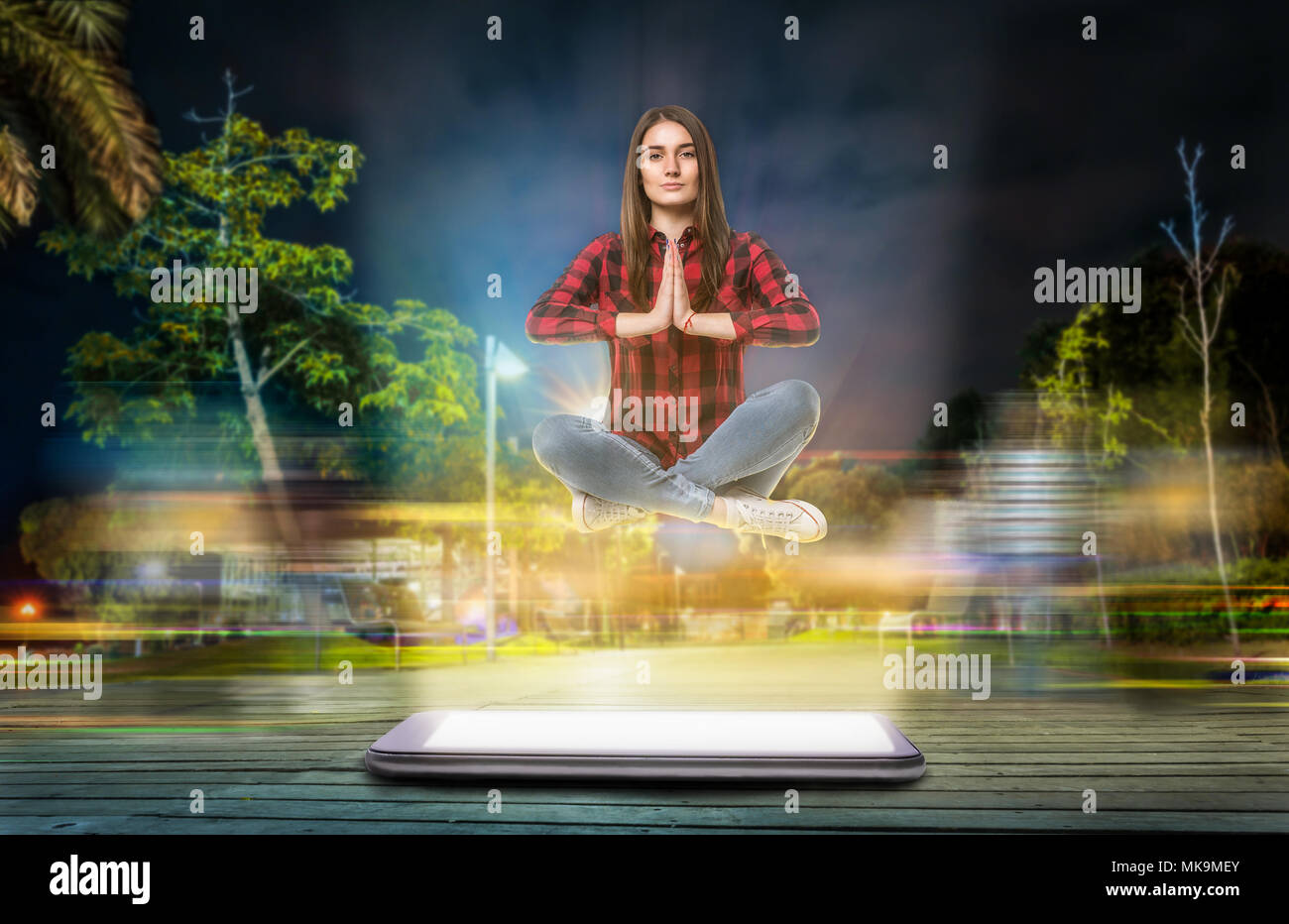 Young woman in yoga pose hovers above a big phone with a glowing screen, evening street with long exposure light effect on background. Communication g Stock Photo