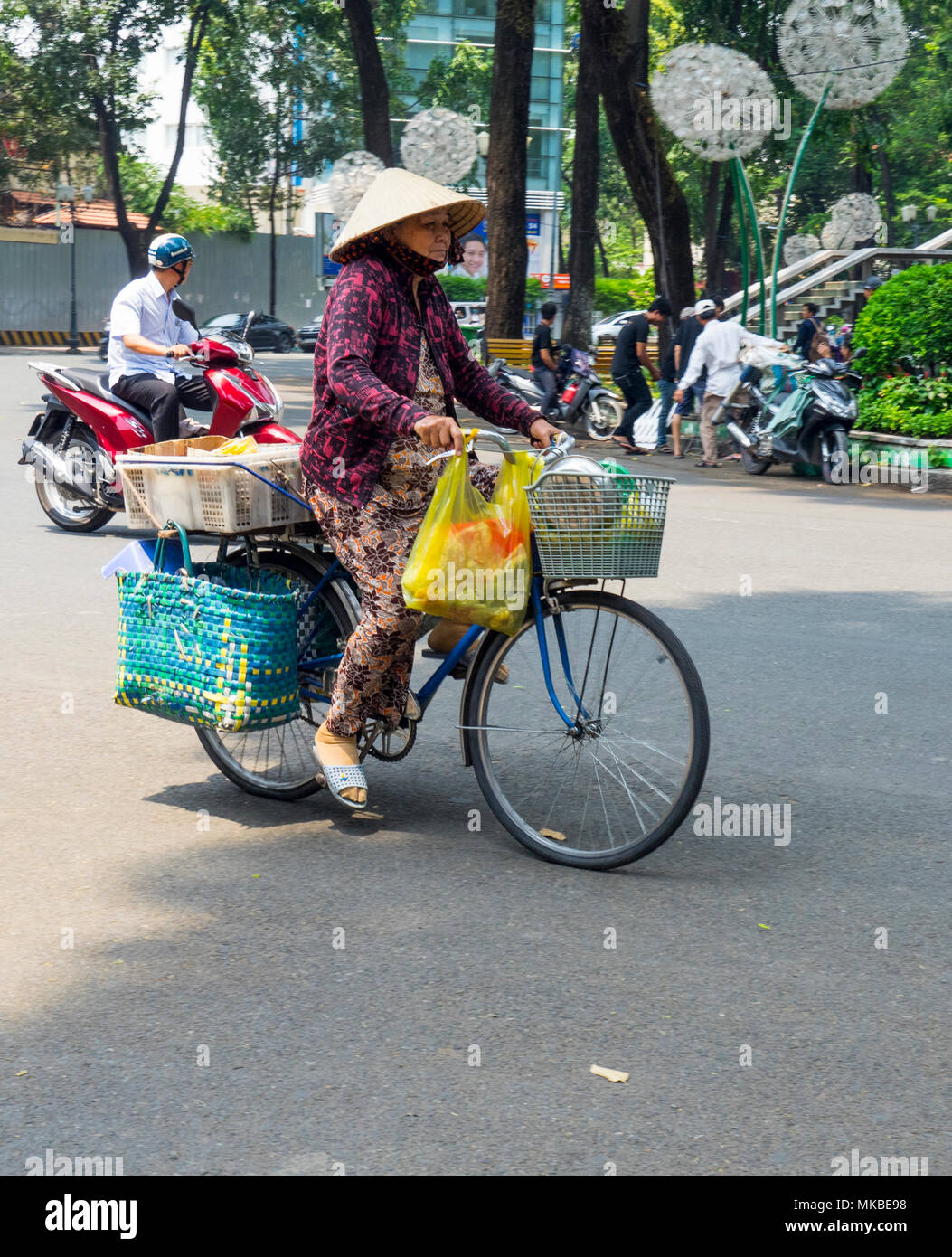 A Vietnamese woman in traditional attire riding a bicycle loaded with her shopping bags in Ho Chi Minh City, Vietnam. Stock Photo