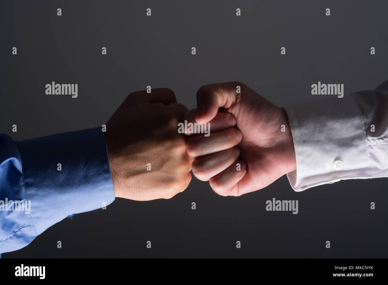 Side view with light source from below of fist bump handshake between businessmen over dark gray background Stock Photo