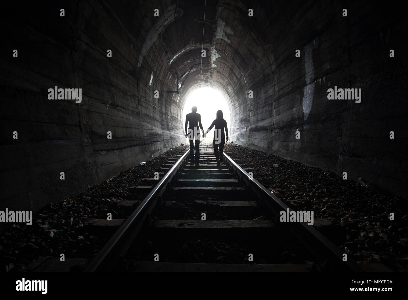 Couple walking hand in hand along the track through a railway tunnel towards the bright light at the other end, they appear as silhouettes against the Stock Photo