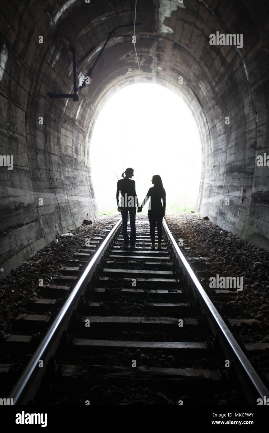 Couple walking hand in hand along the track through a railway tunnel towards the bright light at the other end, they appear as silhouettes against the Stock Photo