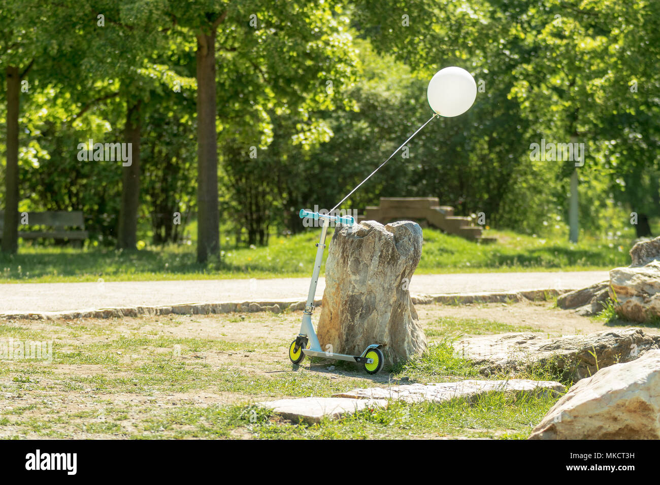 Folding scooter leaning against a large stone in the park. The white balloon filled with helium tied on the handlebars. Lush growing vegetation around Stock Photo