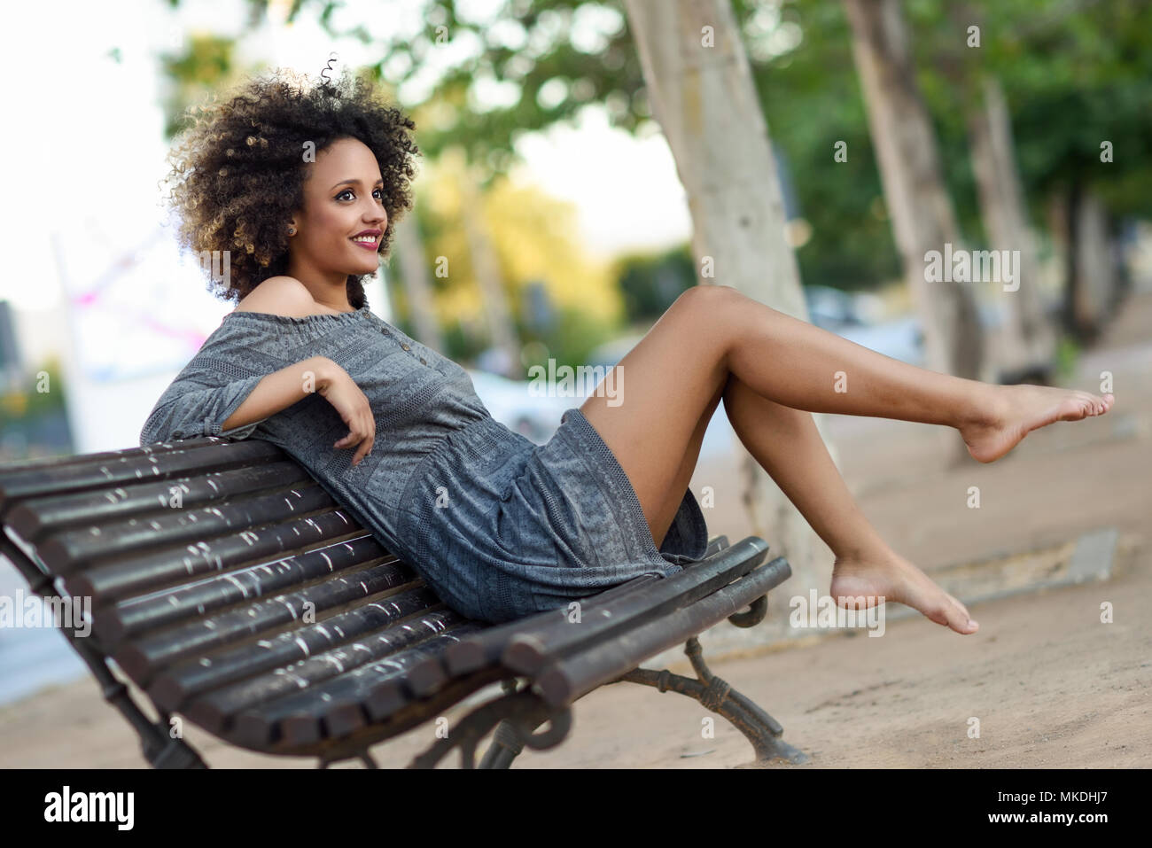 Young black woman with afro hairstyle sitting on a bench in urban background moving her legs. Mixed girl wearing casual clothes. Stock Photo