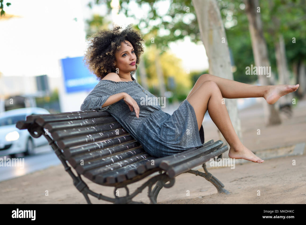Young black woman with afro hairstyle sitting on a bench in urban background moving her legs. Mixed girl wearing casual clothes Stock Photo