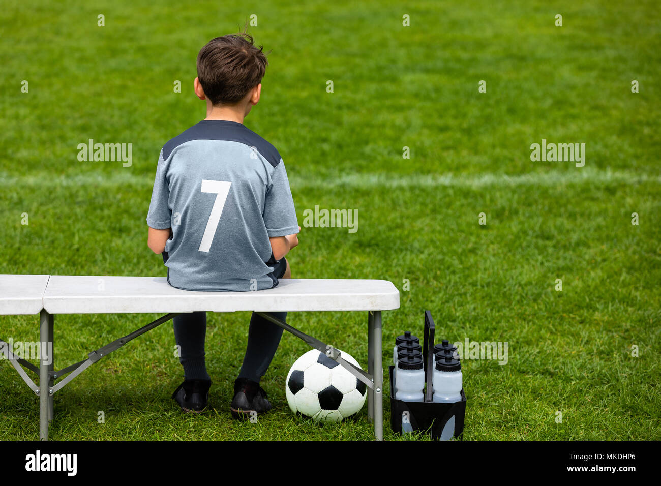 Little footballer sitting on a wooden bench and watching soccer game. Young substitute player waiting on a soccer bench. Boy on a grass pitch with a s Stock Photo