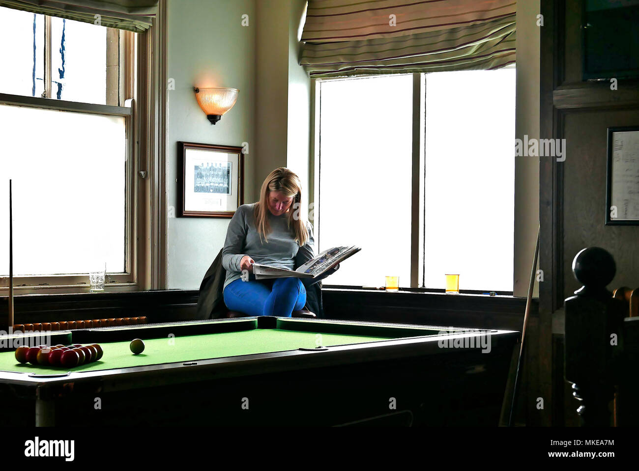 Young women sat on bar stool reading newspaper whist waiting for a game of pool Stock Photo