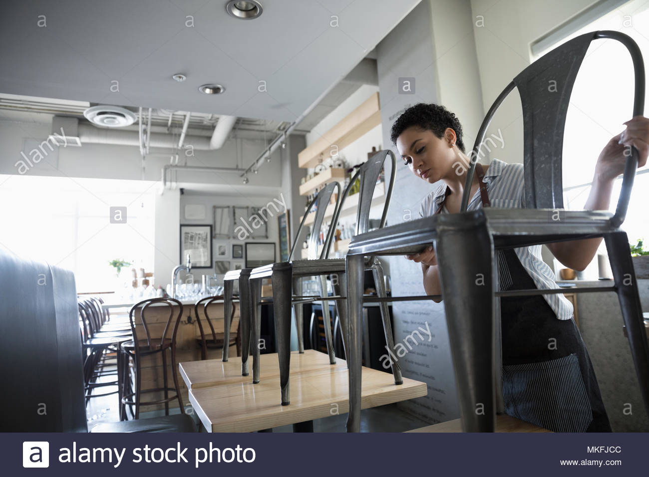 Young woman small business owner stacking chairs on tables in cafe