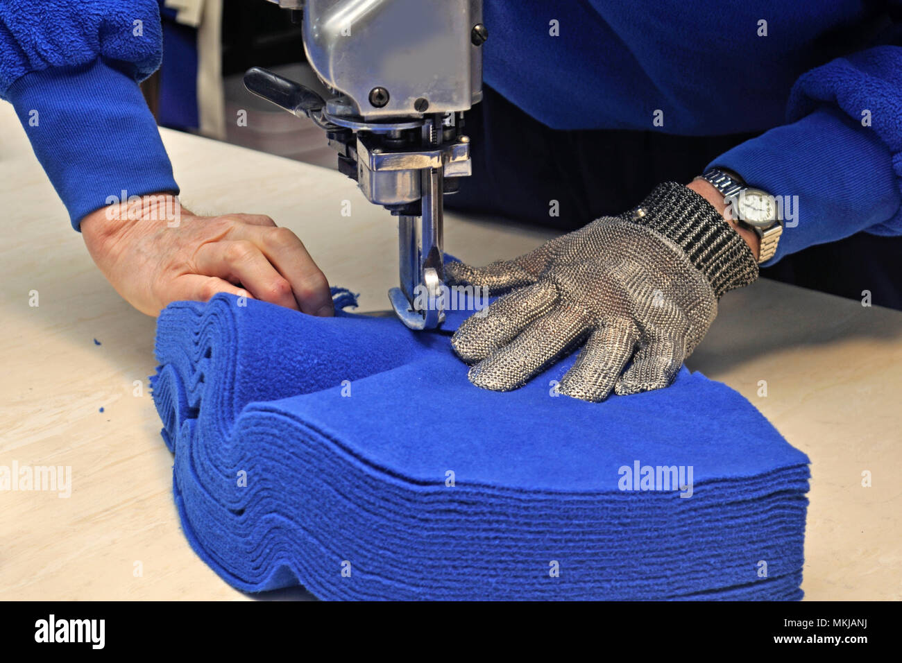 A seamstress uses a vertical knife to cut out a pile of fabric for jackets in a commercial sewing room. Stock Photo
