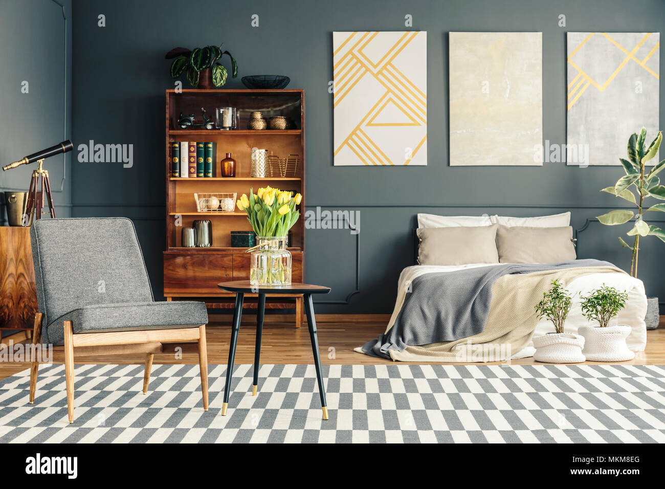 Cozy bed in retro bedroom interior with blankets and cushions next to plants, posters, chair, table, carpet, bookshelf, telescope and books Stock Photo