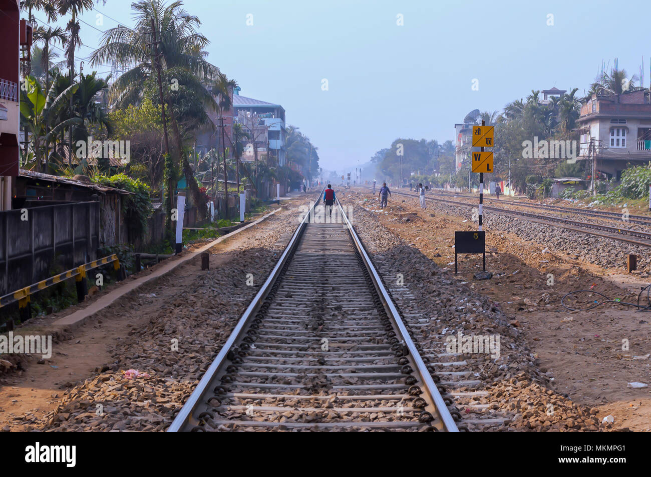 A man walks nonchalantly on the sleepers of a railway tracks, in India, while workers work next to the tracks. Residential houses abut the lines. Stock Photo