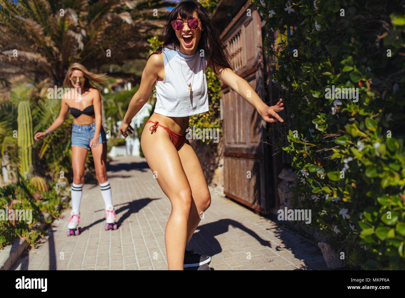 Smiling woman skating during a summer vacation with female friend at the back. Female friends doing skateboarding on the beach. Stock Photo