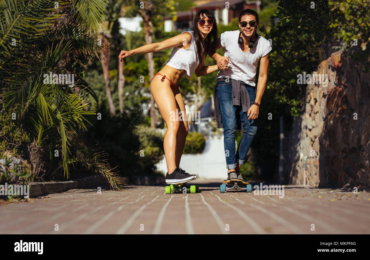 Happy couple riding on skateboards on walkway. Man and woman in summer clothes skating together on vacation. Stock Photo