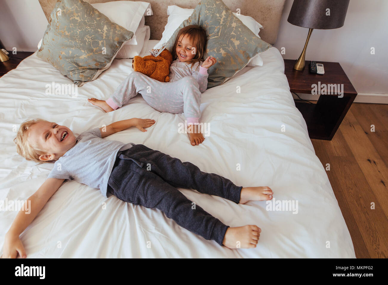 Little boy lying on bed with her sister sitting by holding a teddy bear smiling. Joyful little children playing in bedroom. Stock Photo