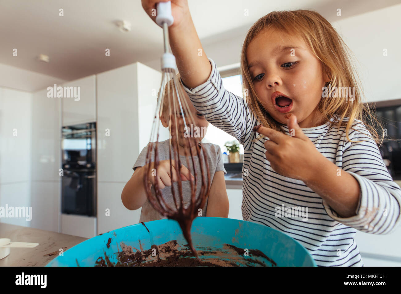 Young girl licking batter from bowl. Little girl mixing the batter with whisk in kitchen. Stock Photo