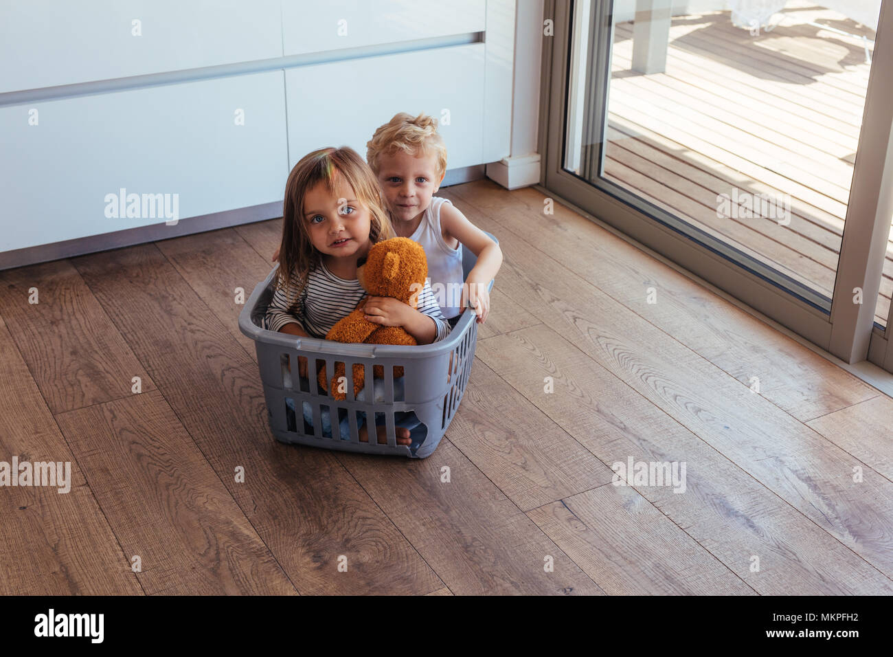 Beautiful young children sitting in a washing basket at home. Little girl holding a teddy bear and her brother playing at home. Stock Photo