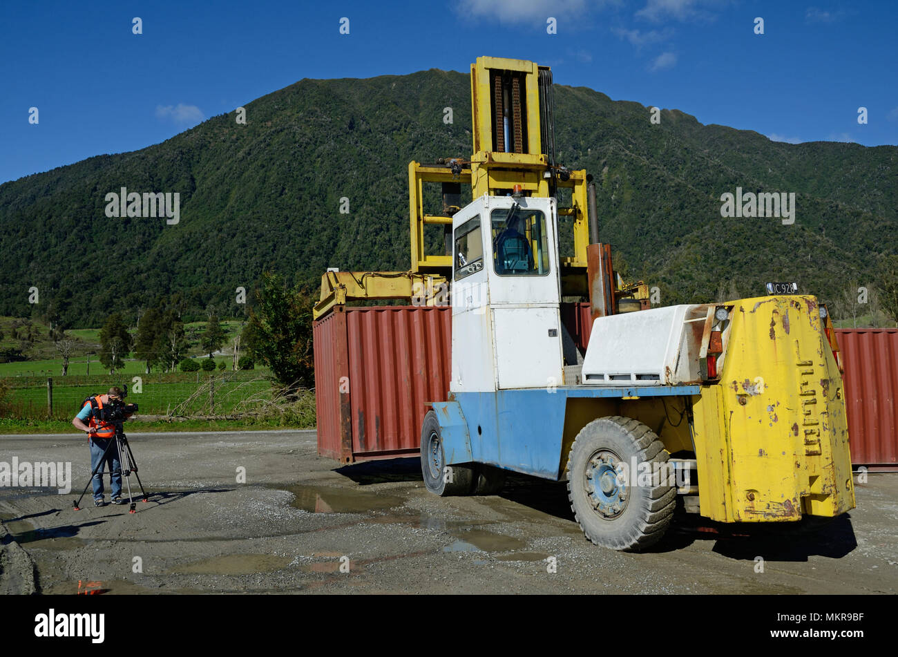 A cameraman films a huge forklift shifting a 20 foot shipping container in the yard of a large factory. Stock Photo