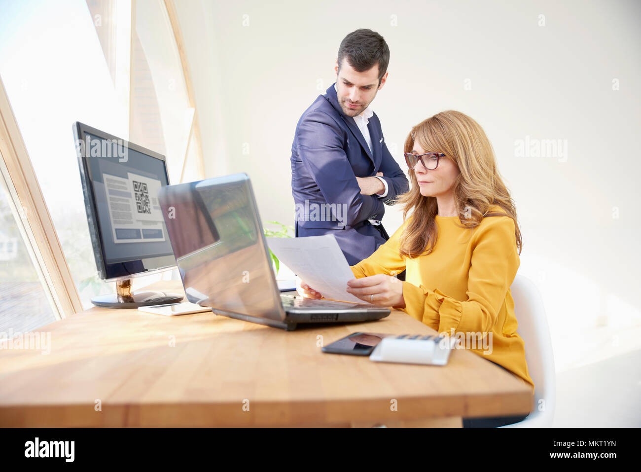 Middle aged businesswoman sitting at office desk and holding a document in her hands while young financial assistant businessman standing next to her  Stock Photo