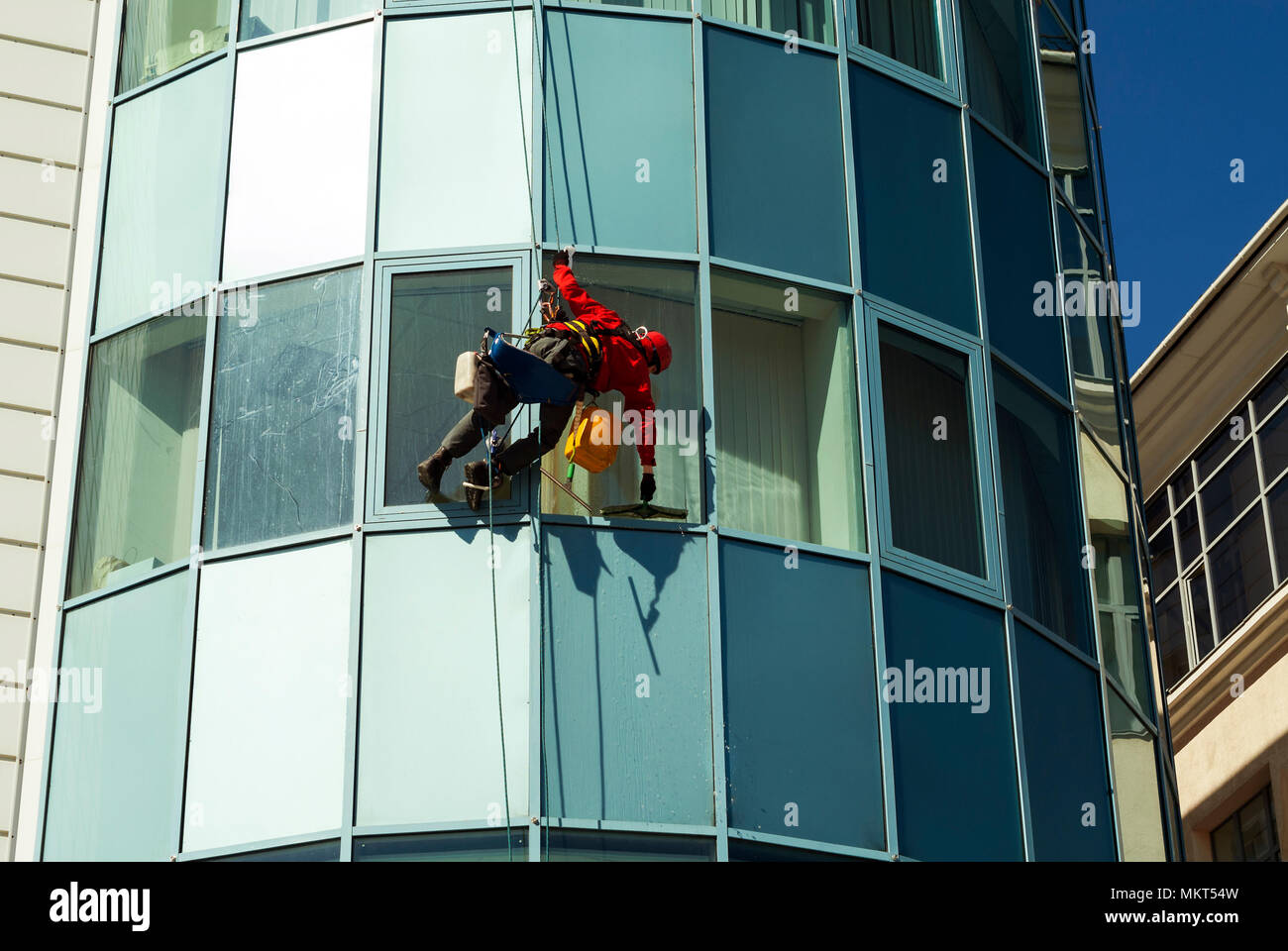 rope access technician in a helmet washes the window of a high-rise building from the outside, hanging on a rope Stock Photo