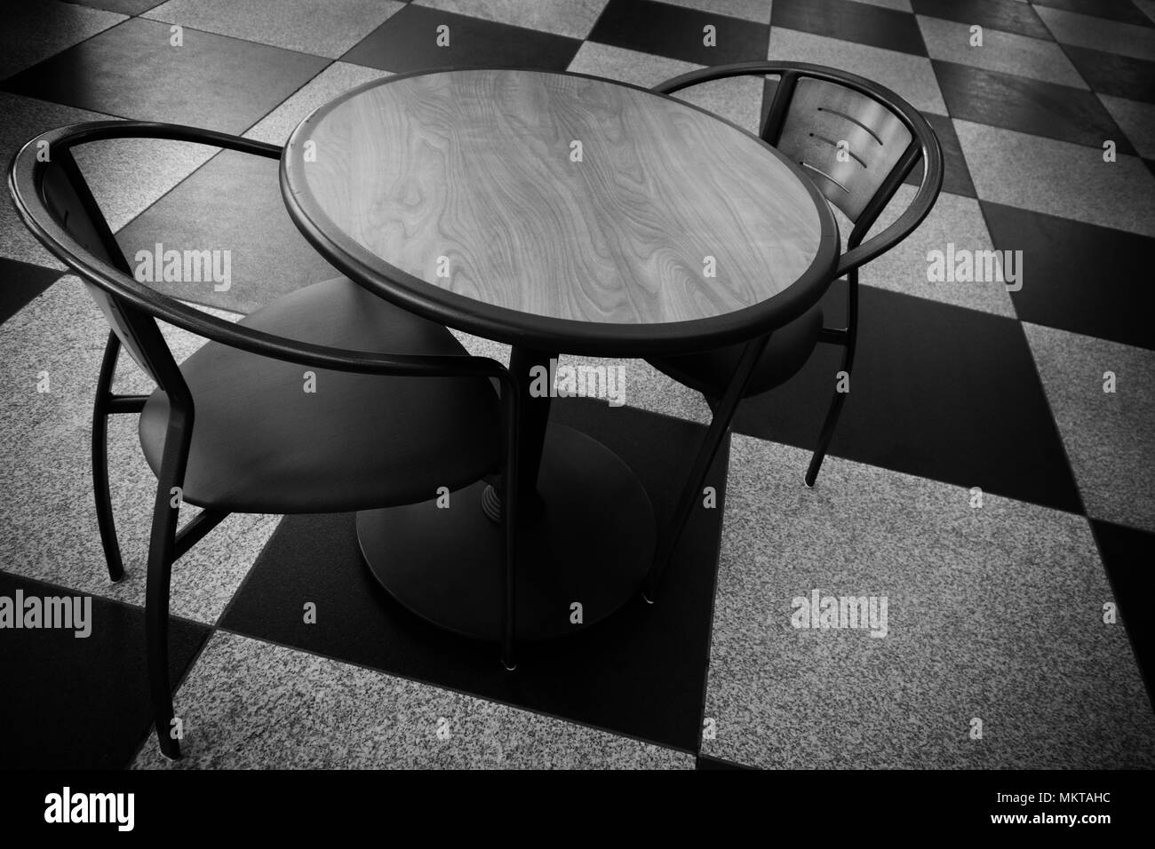 A black and white image of a table and two chairs on a floor with a checkerboard pattern Stock Photo