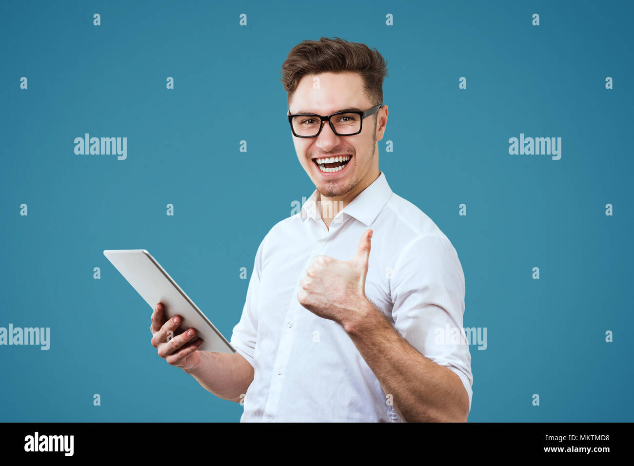 Excited young man wearing white shirt and glasses using tablet pc giving thumbs up gesture isolated on blue background Stock Photo