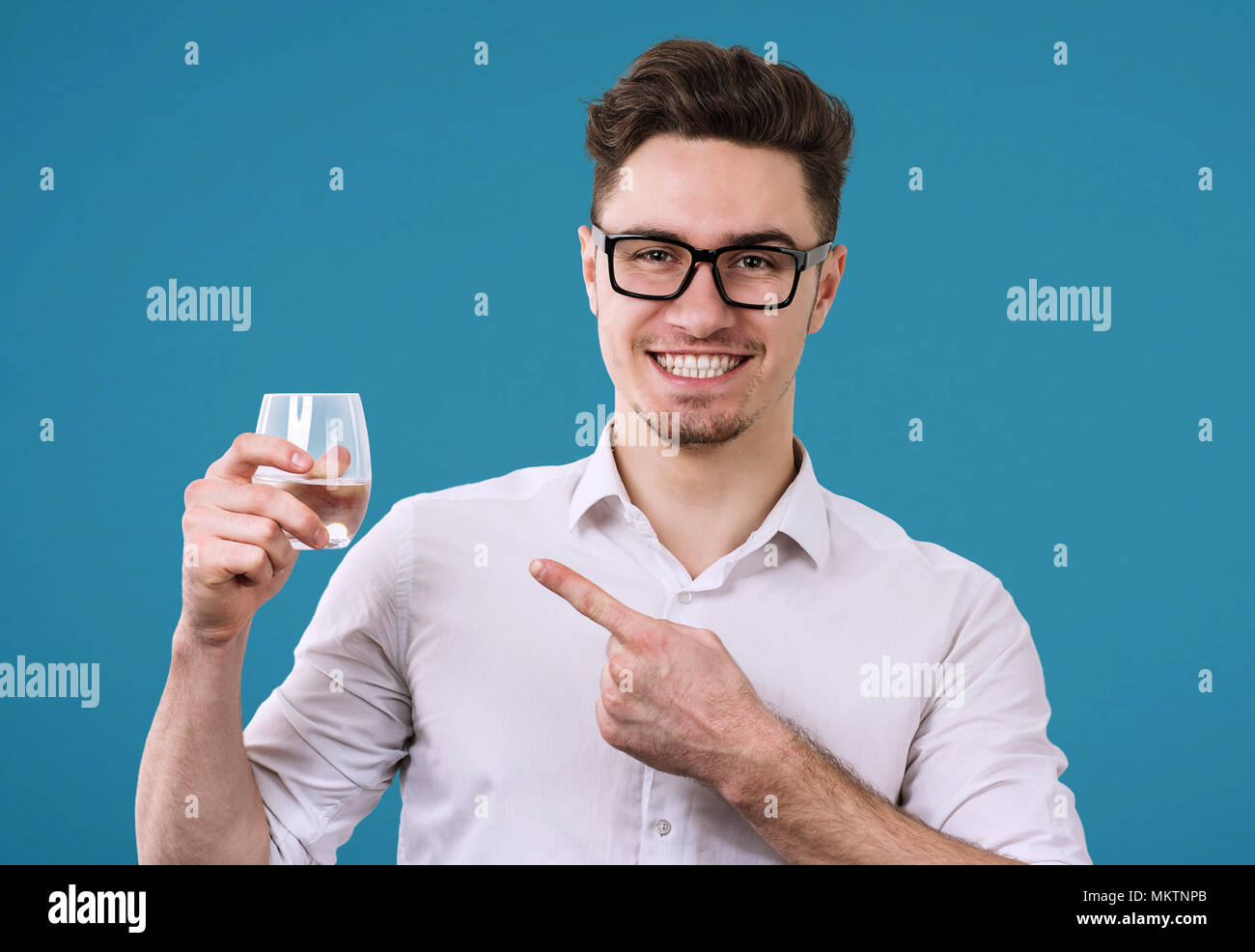 Cheerful handsome man in eyeglasses and holding glass with refreshing drink pointing at it on blue background. Stock Photo