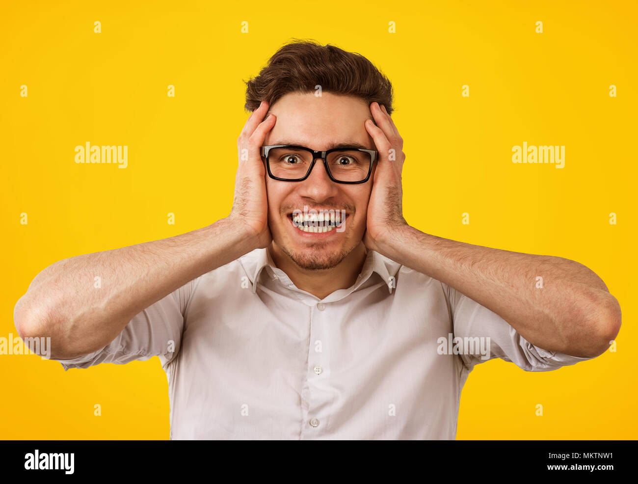 Excited young man in glasses holding hands on head in disbelief looking amazed at camera on yellow background. Stock Photo