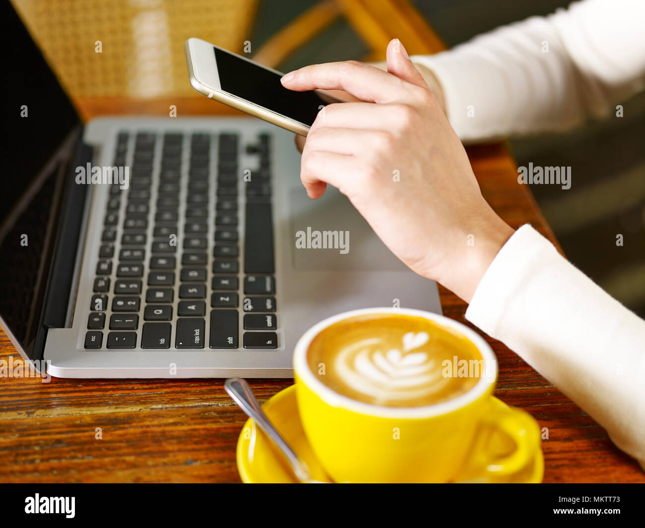 close-up of hands of a woman using mobile phone and laptop computer while drinking cappuccino. Stock Photo
