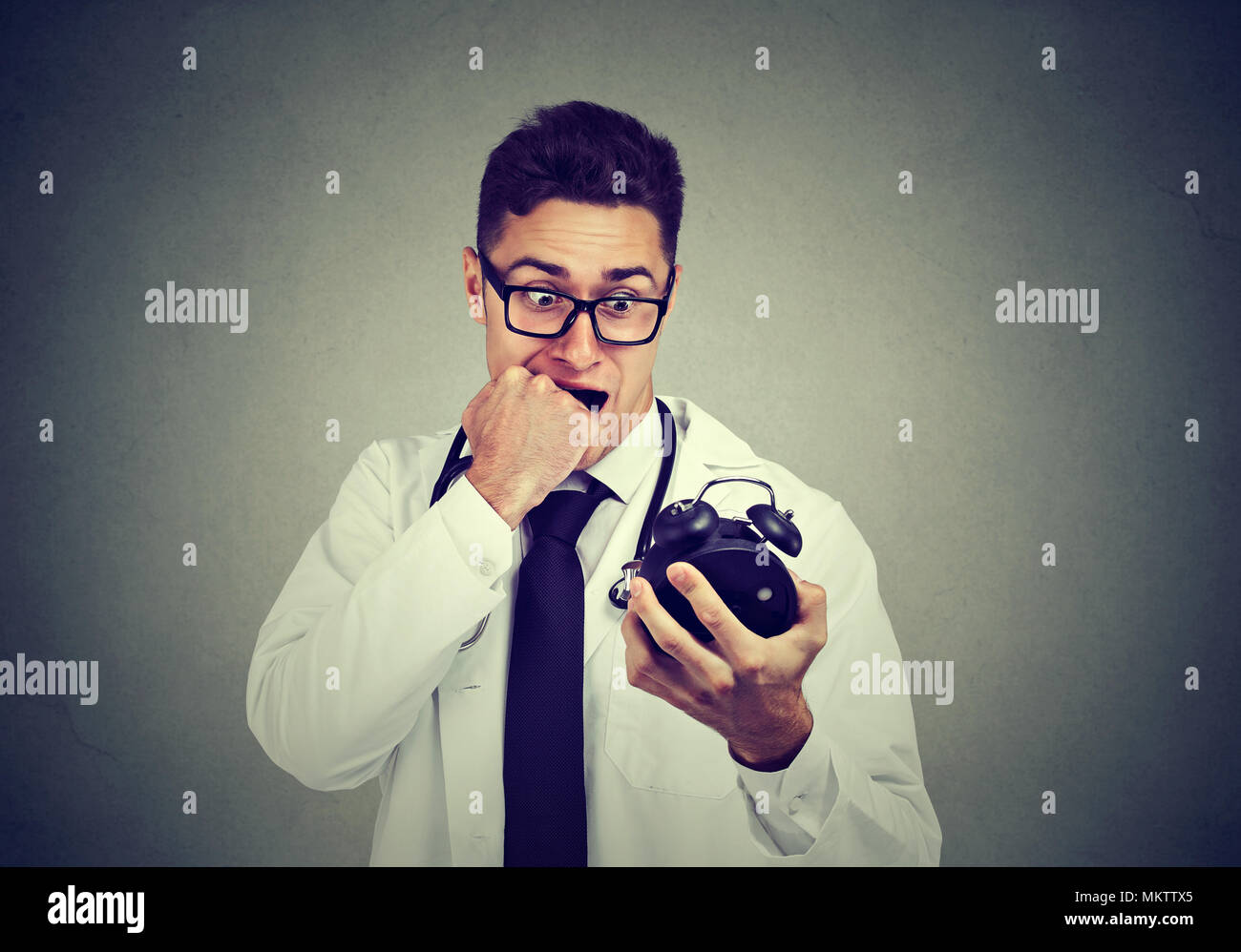 Stressed anxious doctor, health care professional holding alarm clock pressured by time isolated on gray background. Stock Photo