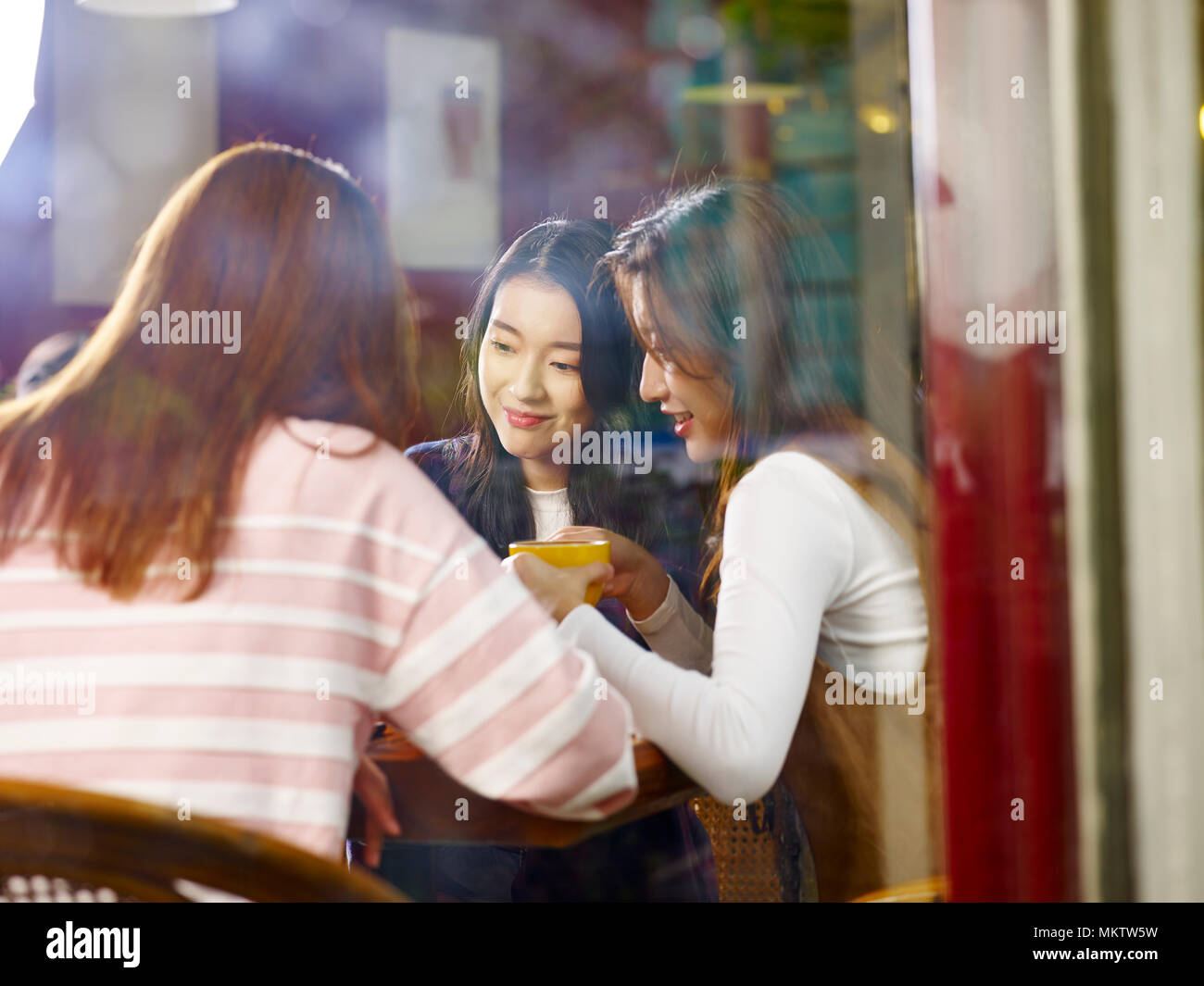 three happy beautiful young asian women sitting at table chatting talking in coffee shop or tea house, shot through window glass. Stock Photo
