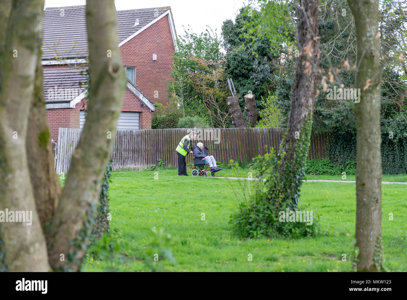 Carer pushes and talks to a man in his wheelchair along the path through Bruche Park, Warrington, Cheshire, England, UK on 29 April 2018 Stock Photo