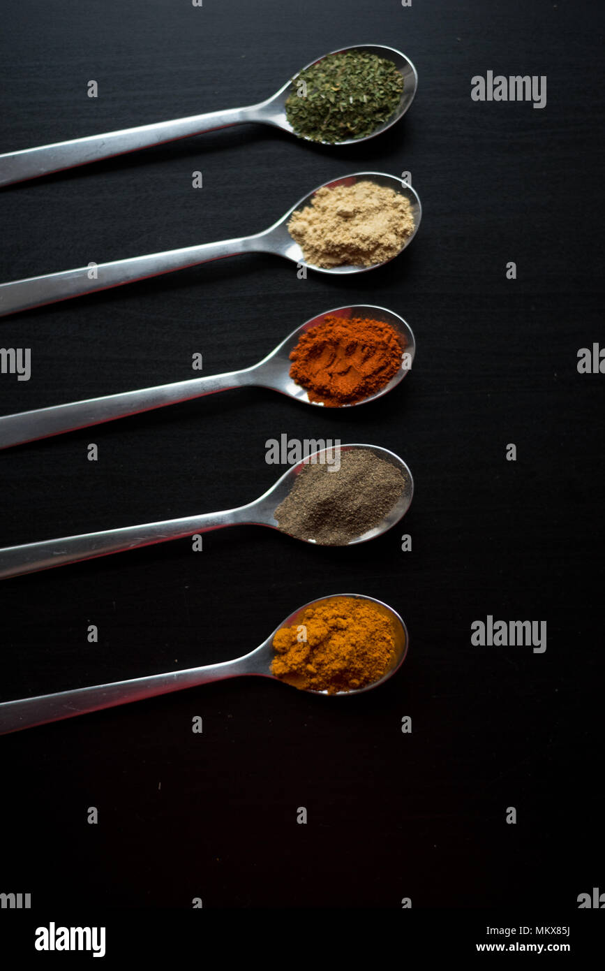 Spices colored in silver spoons, placed on a black wooden table. Pepper, pepper, salt, basil, mustard Stock Photo