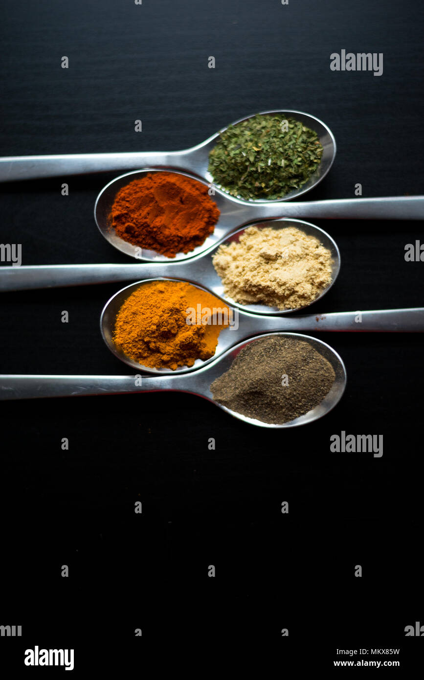 Spices colored in silver spoons, placed on a black wooden table. Pepper, pepper, salt, basil, mustard Stock Photo