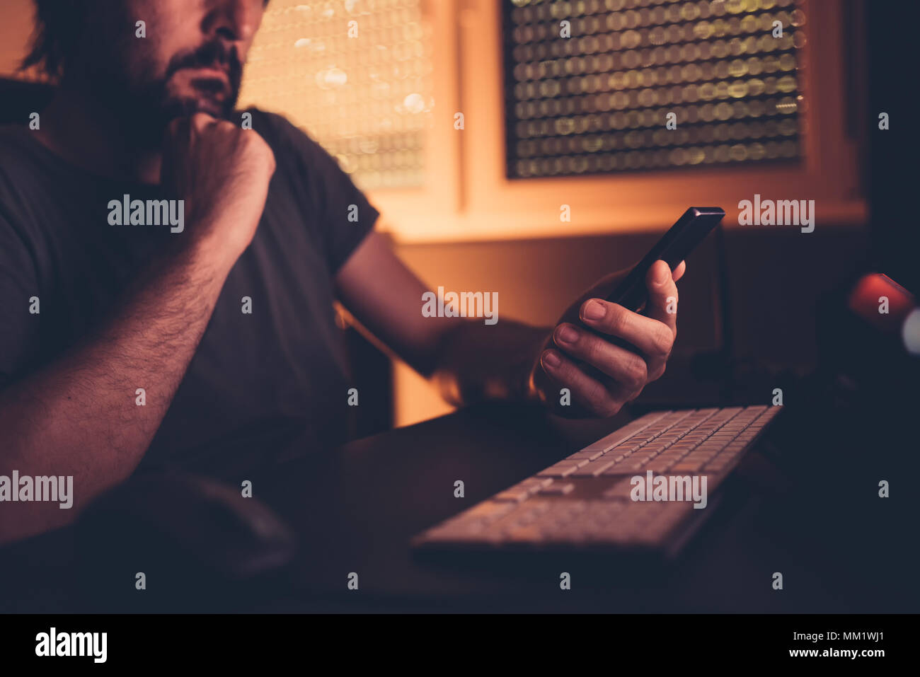 Bored man wasting time using mobile phone in dark home office interior, low key with selective focus Stock Photo