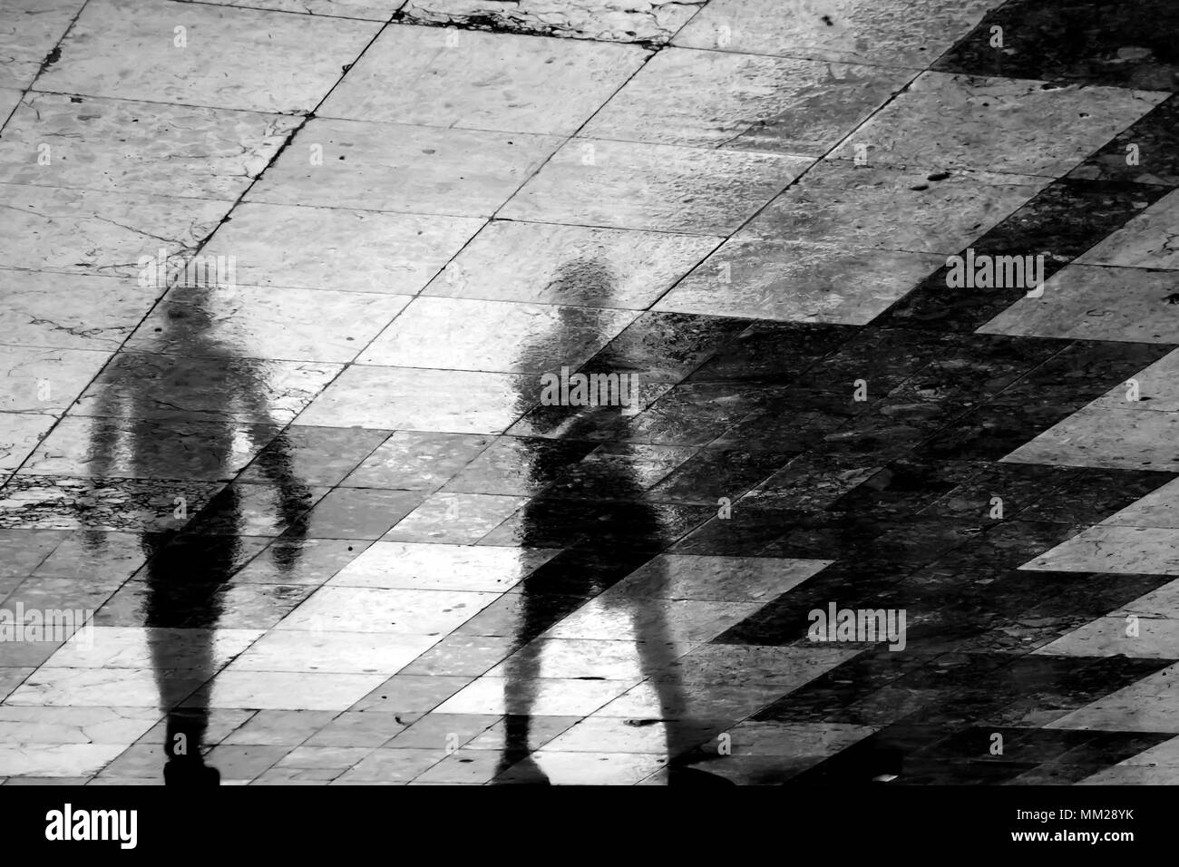 Blurry reflection shadow of two people walking the city street patterned sidewalk on a  rainy spring day Stock Photo
