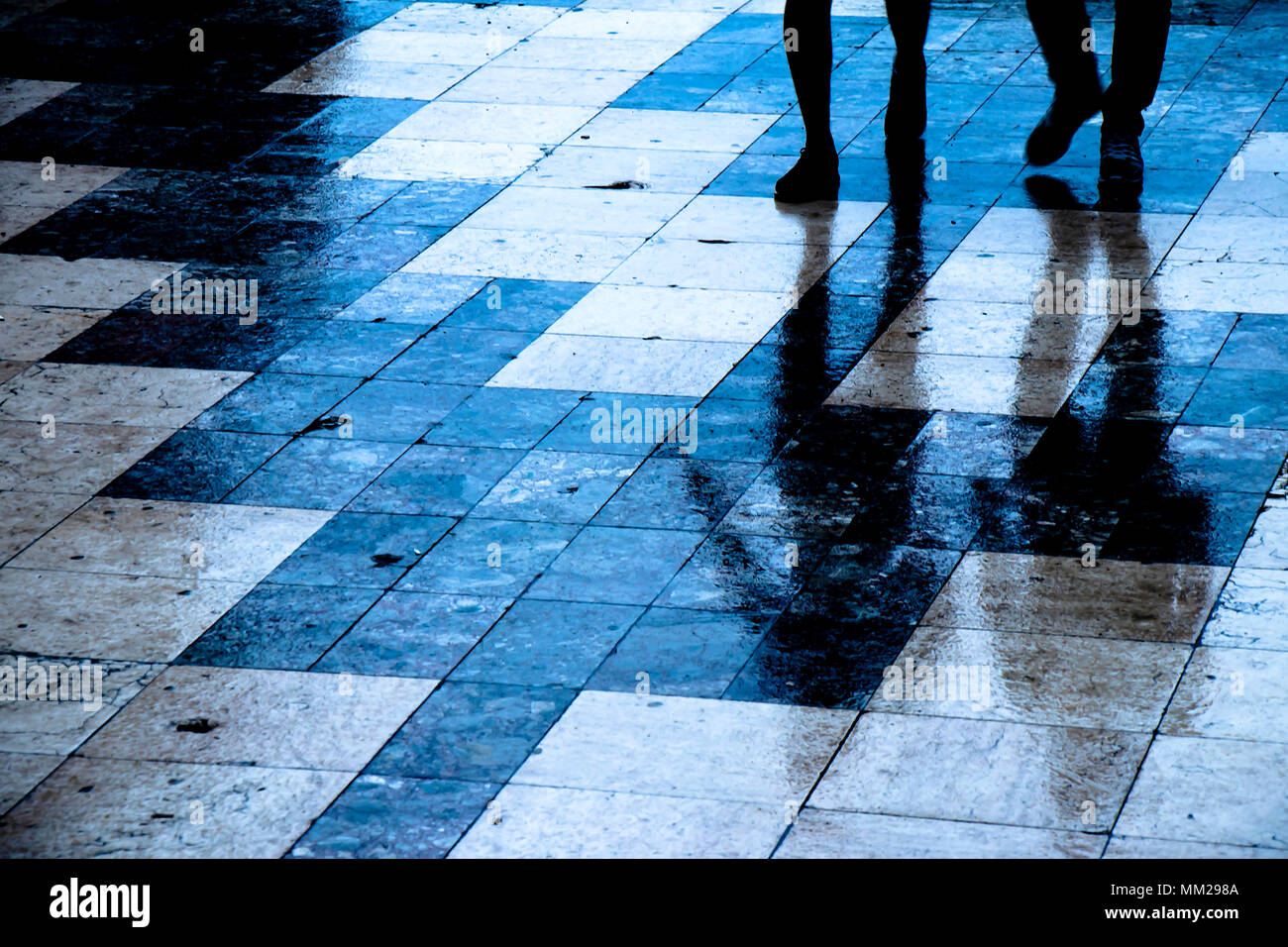 Blurry reflection shadow of two people walking the city street patterned sidewalk on a  rainy night Stock Photo