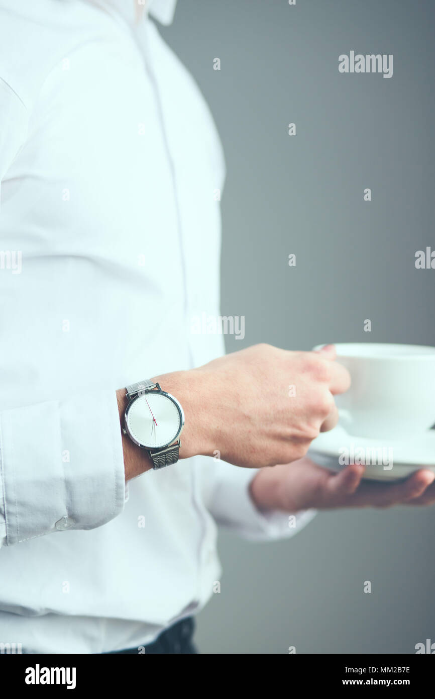 Young man wearing silver wristwatch and white plain shirt holding cup of coffee. Standing in front of plain grey wall Stock Photo