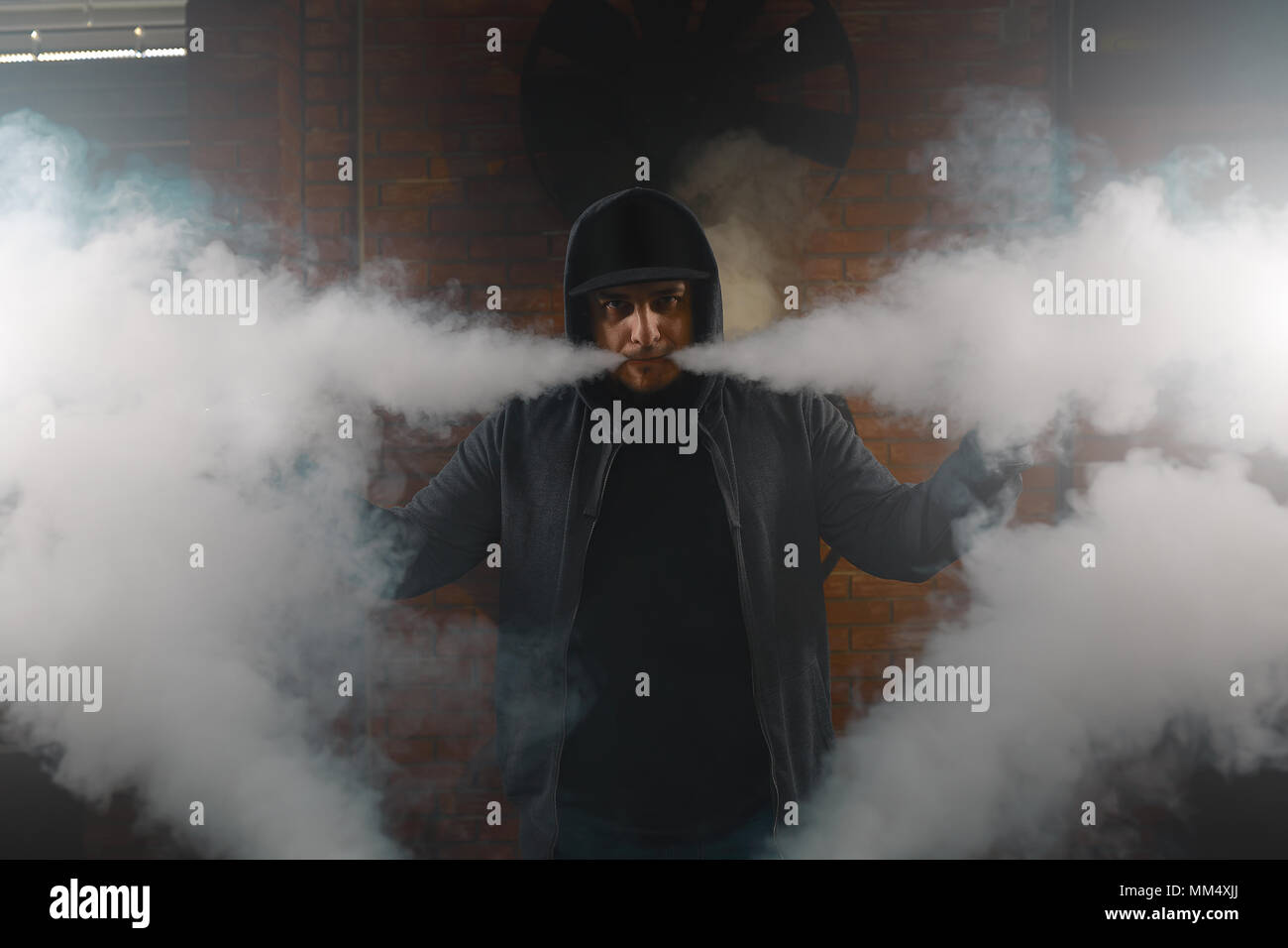 A cloud of vapor. Vaping man holding a mod. Brick background. Vape concept. Young man with beard vaping an electronic cigarette. Vaper hipster smoke v Stock Photo