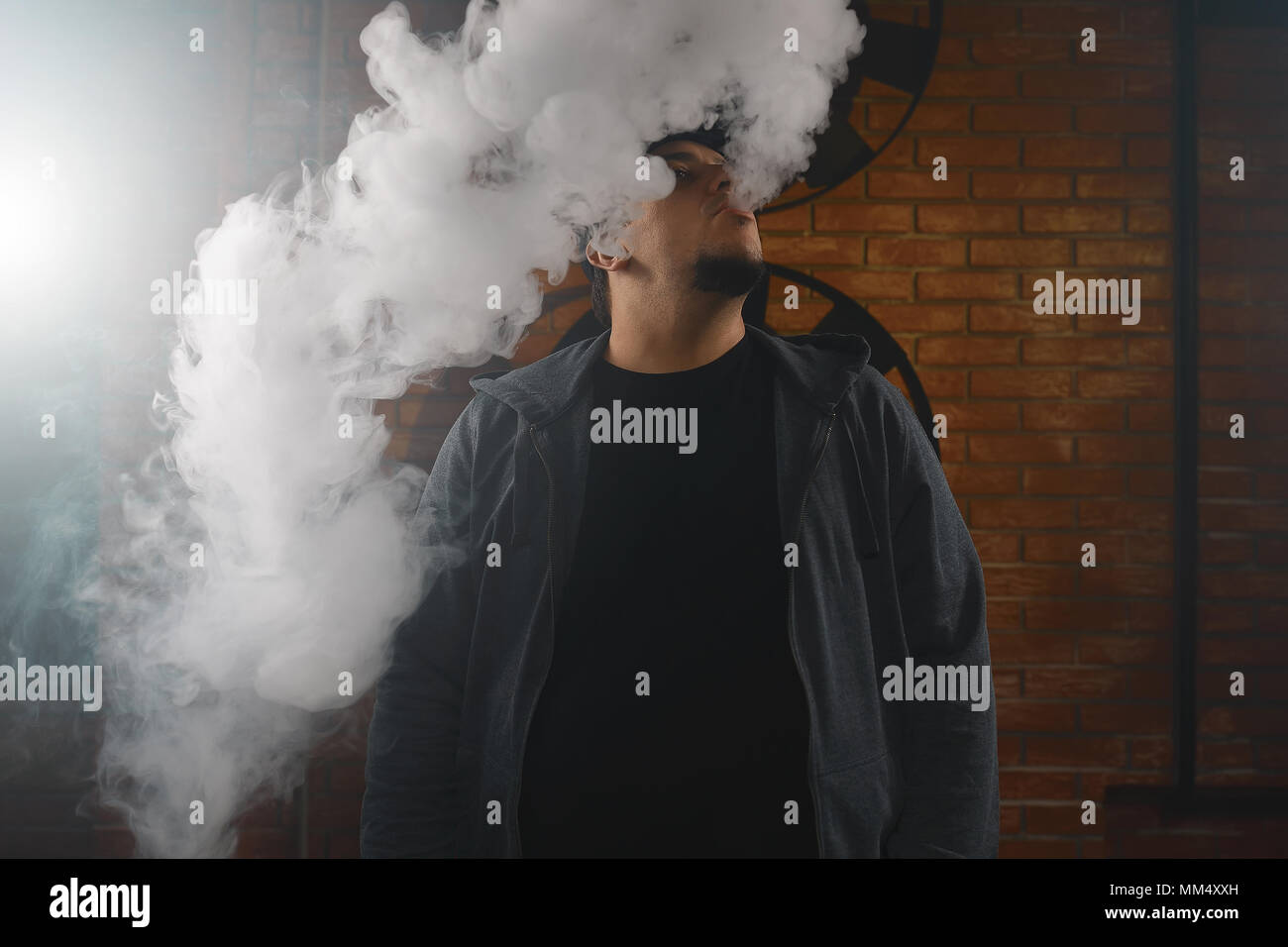 A cloud of vapor. Vaping man holding a mod. Brick background. Vape concept. Young man with beard vaping an electronic cigarette. Vaper hipster smoke v Stock Photo