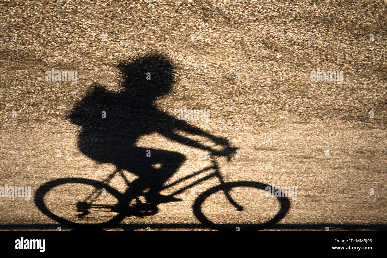 Defocused blurry shadow silhouette of a young person riding a bike on sunset shiny asphalt road Stock Photo