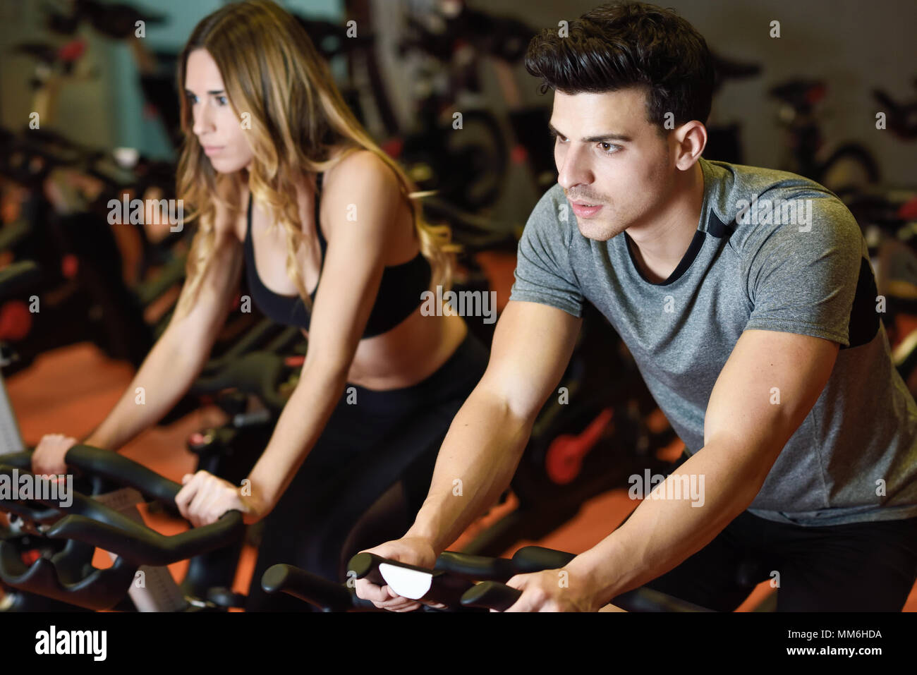Young man and woman biking in the gym, exercising legs doing cardio workout cycling bikes. Two people in a spinning class wearing sportswear. Stock Photo