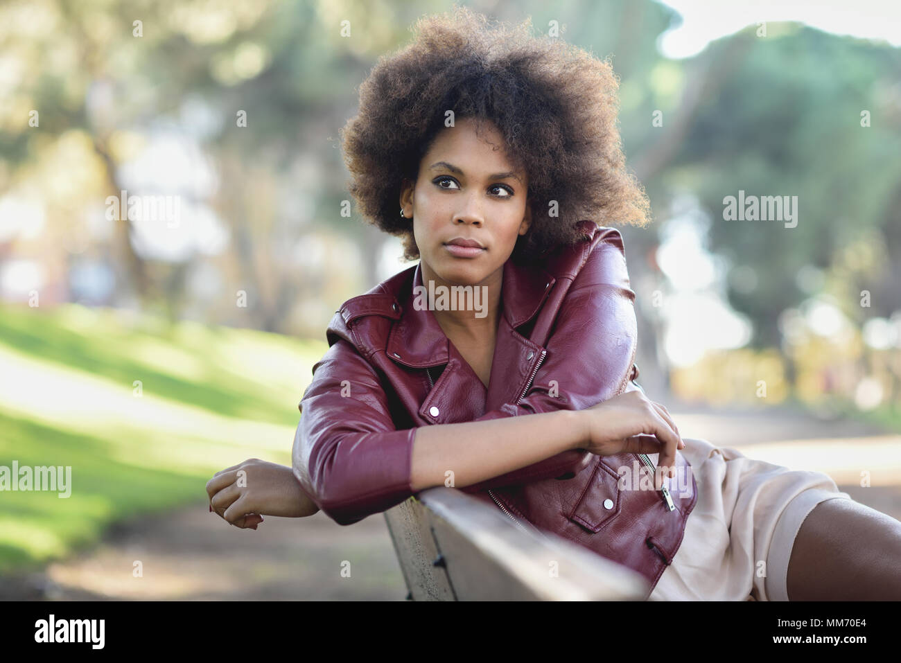 Young black female with afro hairstyle sitting in a bench in an urban park. Mixed woman wearing red leather jacket and white dress with city backgroun Stock Photo