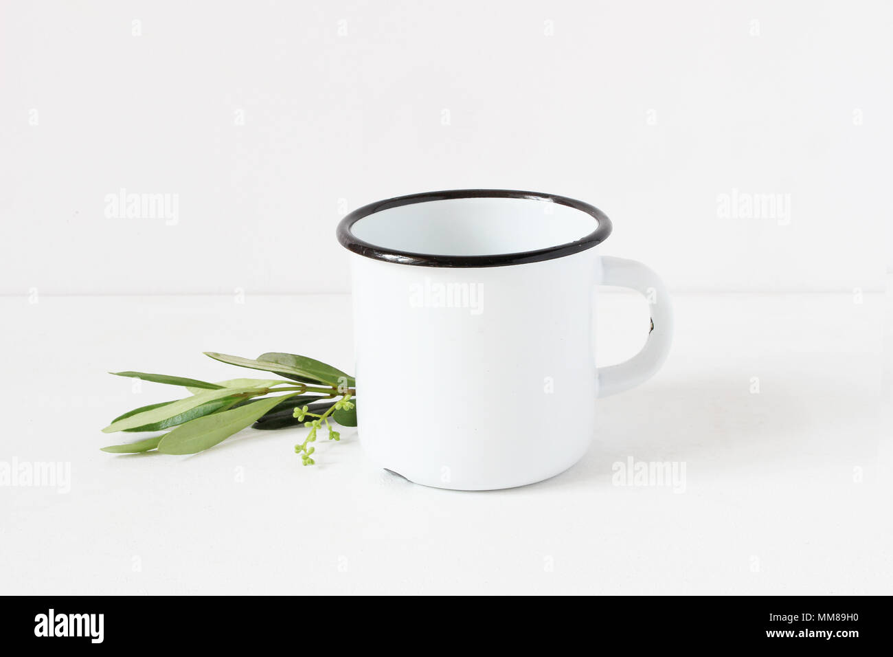 Styled stock photo. Feminine still life composition with blank white metal coffee enamel mug and green olive branch on white table background. Summer rustic scene, product mockup. Stock Photo
